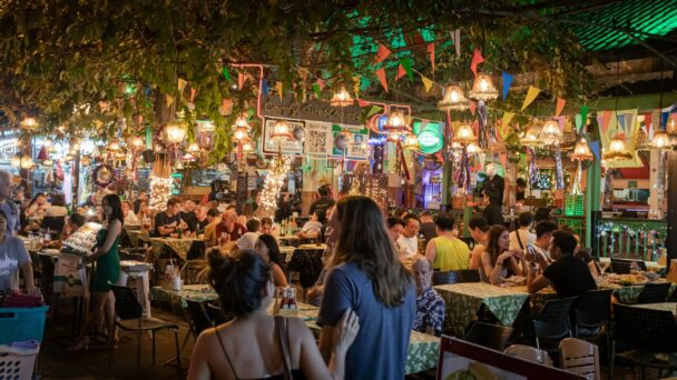 A lively outdoor restaurant is bustling with people dining under twinkling string lights and decorative hanging lamps. Colorful buntings zigzag overhead, and greenery drapes from the ceiling. Patrons sit at closely packed tables, engaged in conversation and enjoying the atmosphere. - Hausion