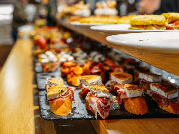 A selection of gourmet tapas on a wooden counter, including small toasted slices of bread topped with various ingredients such as cheese, cured meats, and vegetables. The tapas are arranged on black stone plates with a blurred background. - Hausion