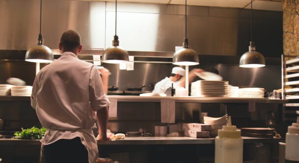 A busy restaurant kitchen with chefs in white uniforms working. In the foreground, a chef is seen from the back organizing ingredients. In the background, other chefs are blurred in motion, plating dishes under industrial lights. Shelves holding plates are visible. - Hausion