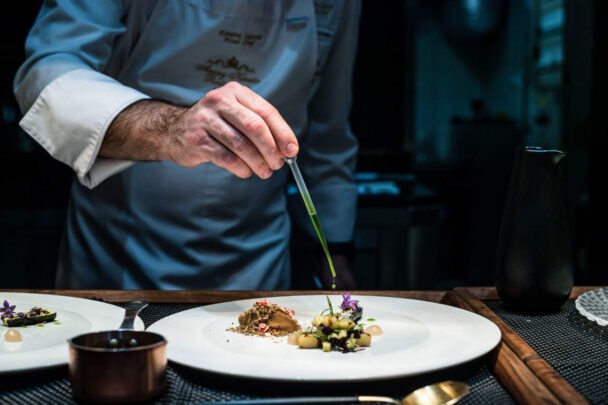 A chef in a white coat carefully garnishes a plated dish with precision using tweezers. The dish is artistically presented with various colorful ingredients on a white plate. A small saucepan and utensils are visible on the table. The setting appears to be a professional kitchen. - Hausion