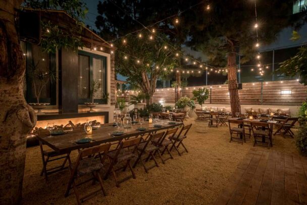 An outdoor dining area is set at night, featuring wooden tables and chairs on a gravel surface. String lights are hung across trees and the surrounding fence, creating a warm, inviting ambiance. The tables are set with plates, glasses, and candles, ready for guests. - Hausion