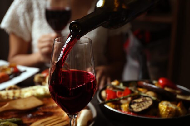 A close-up of red wine being poured into a glass, set against a table filled with various dishes, including grilled vegetables and cheese. The background is slightly blurred, showing a person seated and holding a glass. The ambiance appears warm and cozy. - Hausion