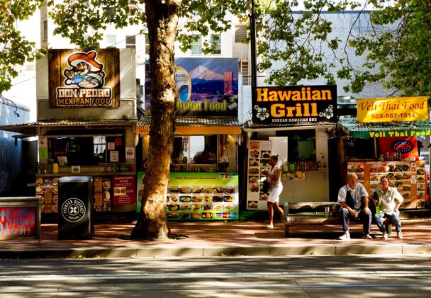 A street scene featuring several food stalls lined up side by side, including "Don Pedro Mexican Food," "Hawaiian Grill," and "Veli Thai Food." Two people sit on a bench in front of the stalls, while two others stand nearby. Trees provide shade over the street and vendors. - Hausion