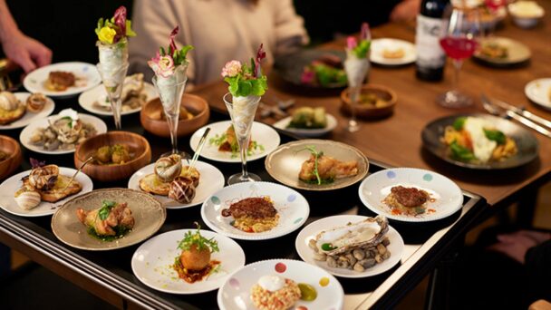 A table is filled with an assortment of elegantly presented dishes, including seafood, meat, and vegetarian options. The plates are meticulously arranged, some with vibrant garnish. Several people are seated around the table, enjoying the meal in a warm, inviting atmosphere. - Hausion