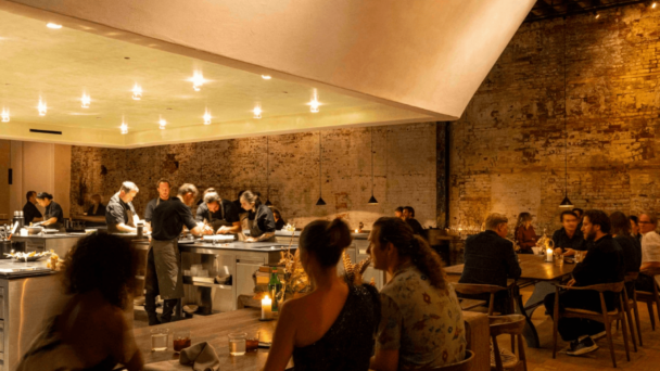 A cozy, dimly-lit restaurant with exposed brick walls. Diners sit at wooden tables, engaged in conversation, while chefs in black attire prepare meals behind a sleek, well-lit counter. Small hanging lights create a warm and intimate ambiance. - Hausion