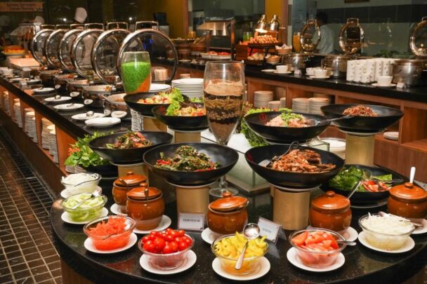 A lavish buffet spread featuring a variety of dishes in large black bowls and jars. The selection includes fresh salads, assorted vegetables, condiments, sauces, meats, and grains. In the background, chafing dishes and plates are arranged for hot items. - Hausion