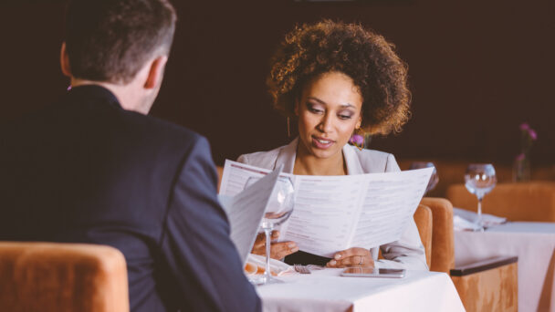 A woman with curly hair and a man in a suit are sitting at a table in a dimly lit restaurant, looking at menus. The table is set with water glasses, a folded napkin, and a smartphone. Red chairs and a dark background are visible. - Hausion