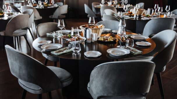 A round dining table set for a meal in an elegant restaurant. The table features several white plates, glasses, napkins, and silverware arranged neatly. A basket of bread and various dishes are placed in the center. Grey cushioned chairs surround the table. - Hausion