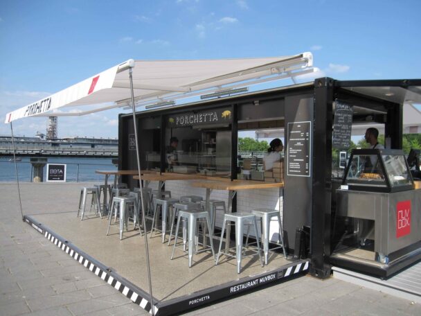 A modern outdoor food stall named "Porchetta" set up in a shipping container, offering shaded bar seating with metal stools. Two staff members are inside, serving food. The stall is located by a waterfront with a view of a bridge in the background. - Hausion