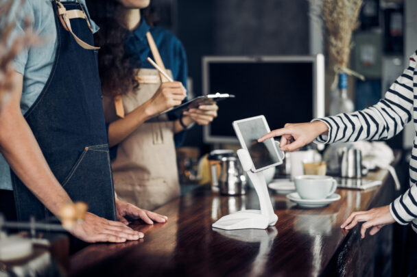 Two cafe staff members, one holding a clipboard, stand behind a wooden counter. A customer is using a touchscreen device on the counter. Drinkware, such as cups and saucers, are visible in the background. - Hausion