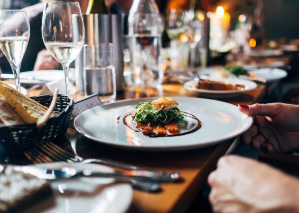 A close-up of a gourmet meal served on a white plate, featuring a small portion of food garnished with greens and sauce. The table is elegantly set with wine glasses, candles, and silverware, suggesting a fine dining experience. - Hausion