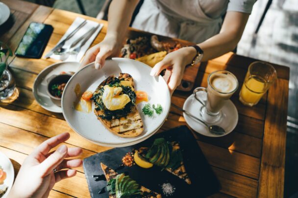 A person hands a plate of toast topped with poached eggs, greens, and tomatoes to another person's outstretched hand over a wooden table. The table is set with various dishes, including a latte, a glass of orange juice, and avocado toast. - Hausion