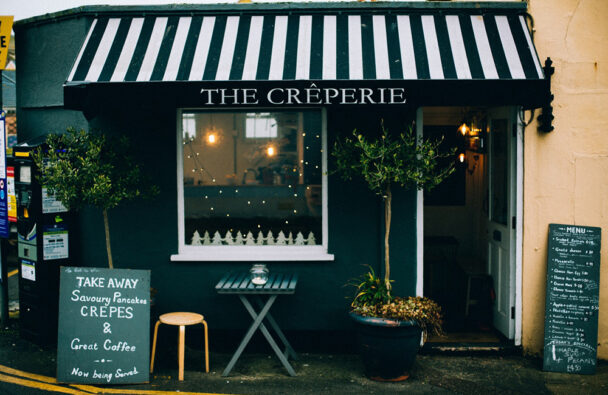 A cozy crepe cafe with a black and white striped awning labeled "The Crêperie." Outside, there's a chalkboard menu on a small wooden stand, a table with two chairs, potted plants, and festive lights around the window. A larger menu is on the right wall. - Hausion