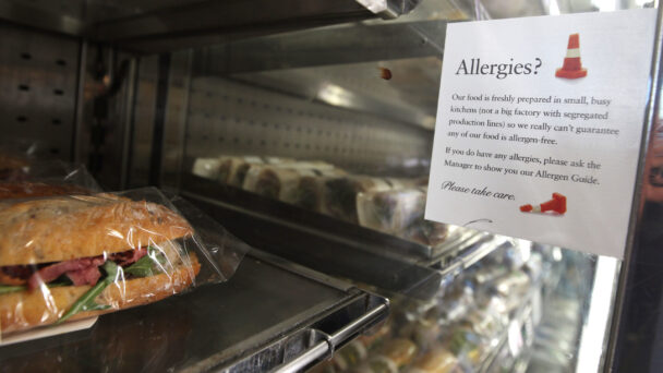 A display case with sandwiches is shown. Next to the sandwiches, there is a sign that reads, "Allergies? Our food is freshly prepared in small, busy kitchens (not in a factory with segregated production lines) so we really can't guarantee any of our food is allergen-free. - Hausion