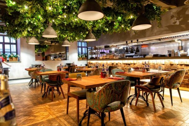 A cozy restaurant with wooden tables and a variety of patterned chairs. The ceiling is adorned with hanging greenery and pendant lights, creating a warm, inviting atmosphere. In the background, there is an open kitchen with chefs preparing meals. - Hausion