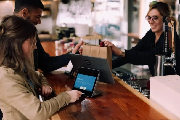 A barista hands a paper bag to a customer across the counter in a café. The customer is making a payment on a digital point-of-sale terminal, while another customer stands beside them. The scene shows a friendly transaction in a well-lit establishment. - Hausion