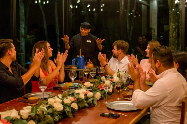 A lively group of friends at a table, hands raised in excitement, sharing joyful moments together. - Hausion