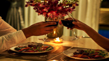 A person arranging a beautiful flower bouquet in a glass vase with a candle and a meal set up for a romantic dinner with a glass of red wine.