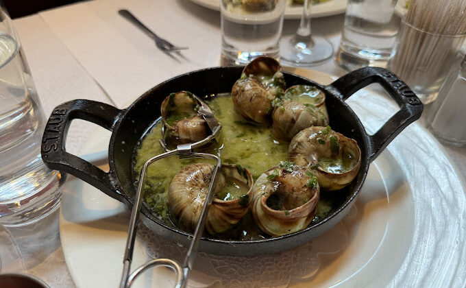 A dish of escargot served in a black cast-iron pan, featuring several cooked snails in shells swimming in a garlic and herb butter sauce. A special snail tong is placed on the edge of the pan. The table includes water glasses, cutlery, and other dishes in the background. - Hausion