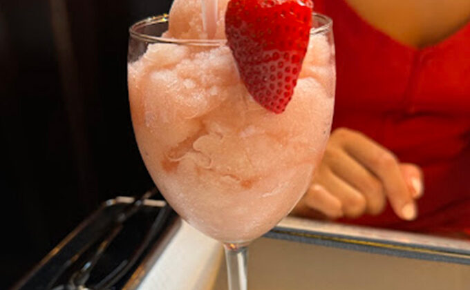 A glass of pink frozen beverage with a fresh strawberry garnish, placed on a table. Behind the glass, part of a person wearing a red shirt is visible. The blurred background suggests an indoor setting. - Hausion