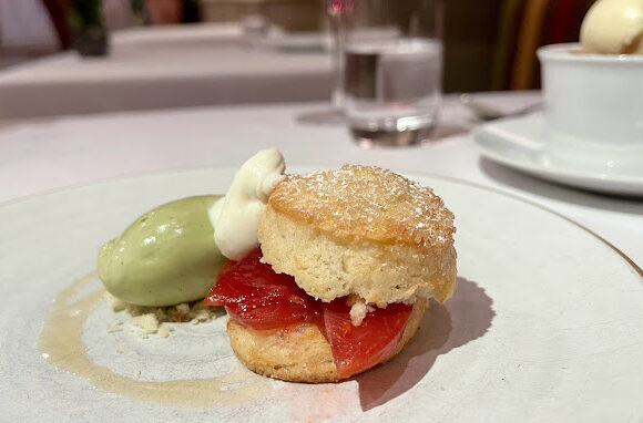 A dessert plate featuring a biscuit topped with a slice of fruit and a dollop of cream, accompanied by a scoop of green ice cream. In the background, there is a glass of water and another bowl with a scoop of vanilla ice cream on a white tablecloth. - Hausion