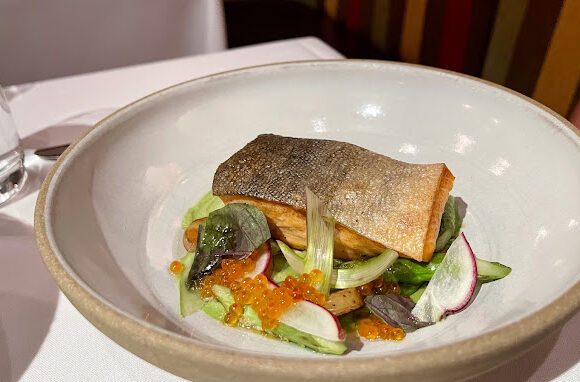 A carefully plated dish featuring a fillet of seared fish with crispy skin, resting on a bed of mixed greens, radishes, and herbs, garnished with orange fish roe. A glass of water is visible to the left side of the white tablecloth. - Hausion