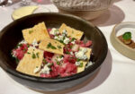 A gourmet dish is presented in a dark bowl, containing finely chopped, seasoned raw meat pieces, crumbled blue cheese, and scattered fresh herbs, topped with thin, crispy bread slices. A dish with small appetizers and a dipping sauce is in the background. - Hausion