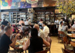 A bustling restaurant interior with patrons seated at wooden tables enjoying their meals. There is a bar in the background with a wide selection of bottles. The walls feature colorful artwork and a large vase with sunflowers decorates the right side of the image. - Hausion