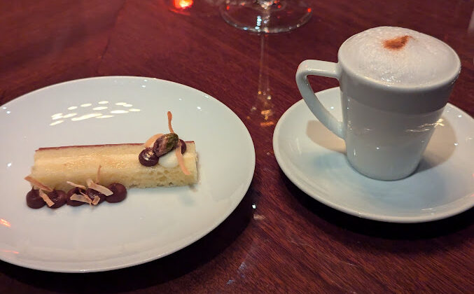 A white plate holds a rectangular dessert garnished with chocolate and nuts, next to a white cup of frothy cappuccino on a saucer, placed on a glossy wooden table. - Hausion