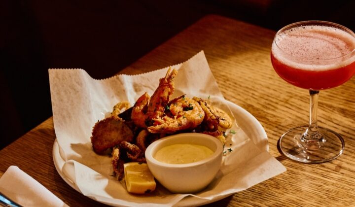 A plate of assorted fried seafood including shrimp, accompanied by a small bowl of dipping sauce and a wedge of lemon, sits on a wooden table. Beside the plate is a pink cocktail in a coupe glass. Napkins, cutlery, and another plate are also visible. - Hausion