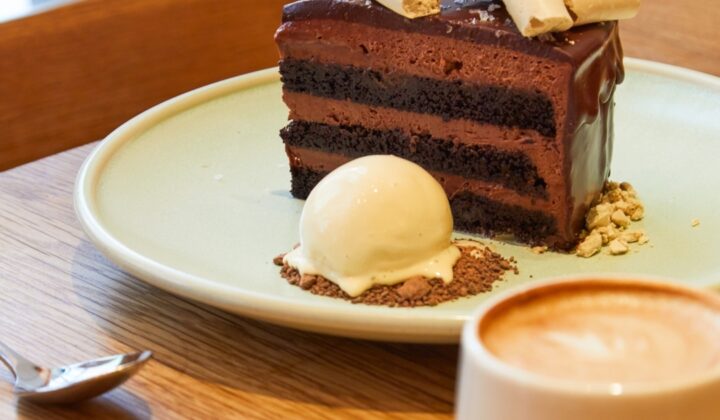 A slice of chocolate layer cake with a smooth chocolate glaze, topped with thin white sticks, is served on a green plate. The plate also holds a scoop of vanilla ice cream garnished with crumbs. Next to the plate is a cappuccino in a white cup with a spoon beside it. - Hausion