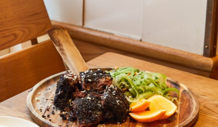 A wooden table set for a meal with a large, rustic wooden platter of barbecued meat topped with seasoning, alongside sliced oranges and a green vegetable side. In the foreground, there are place settings with plates, napkins, and cutlery. - Hausion