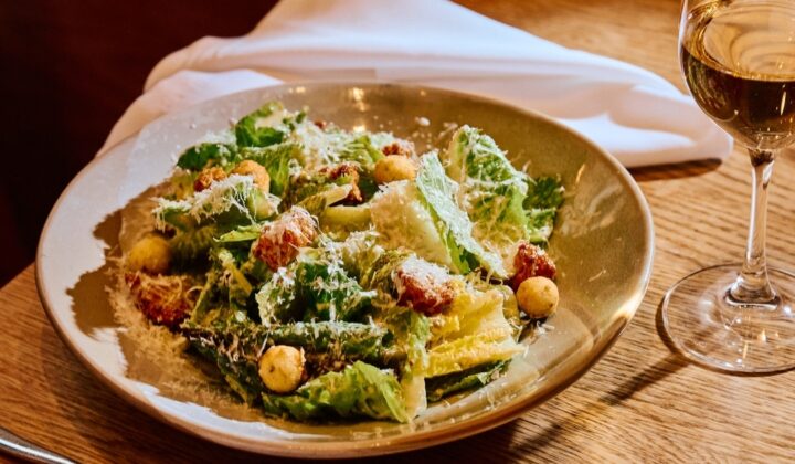 A plate of Caesar salad, topped with croutons and grated cheese, is set on a wooden table beside a glass of white wine. A fork, knife, and white napkin are placed to the left. The background includes part of a chair and warm, ambient lighting. - Hausion