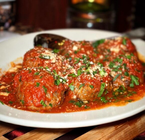 A white plate filled with meatballs covered in tomato sauce, garnished with chopped parsley and grated parmesan cheese. A spoon rests in the background on the plate. The dish is situated on a wooden surface. - Hausion