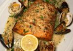 A dish featuring a roasted salmon fillet garnished with chopped herbs, served with clams and mussels in a creamy sauce. The plate is adorned with a halved lemon in the foreground. The seafood is displayed around the salmon. - Hausion