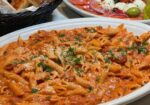 A delicious spread of Italian cuisine features a large oval dish filled with penne pasta in a rich, creamy tomato sauce, garnished with fresh herbs. In the background, there's a basket of sliced bread and a platter with sliced tomatoes, mozzarella, and olives. - Hausion