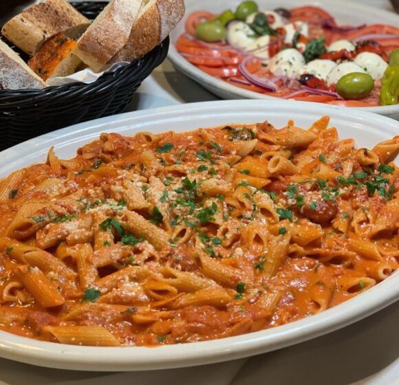 A delicious spread of Italian cuisine features a large oval dish filled with penne pasta in a rich, creamy tomato sauce, garnished with fresh herbs. In the background, there's a basket of sliced bread and a platter with sliced tomatoes, mozzarella, and olives. - Hausion