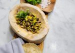 A wooden serving board holds a rustic bowl with grilled vegetables, a small plate with finely chopped onions and greens with an orange flower, a bowl of seasoning, and a neatly wrapped set of tortillas. The board rests on a light marble surface. - Hausion