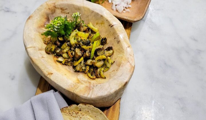 A wooden serving board holds a rustic bowl with grilled vegetables, a small plate with finely chopped onions and greens with an orange flower, a bowl of seasoning, and a neatly wrapped set of tortillas. The board rests on a light marble surface. - Hausion