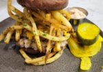 A hearty burger is topped with a generous portion of French fries and melted cheese, served on a round, speckled plate. On the side, there are pickle slices and a small metal cup filled with a creamy dipping sauce. The dish is arranged on a white marble surface. - Hausion