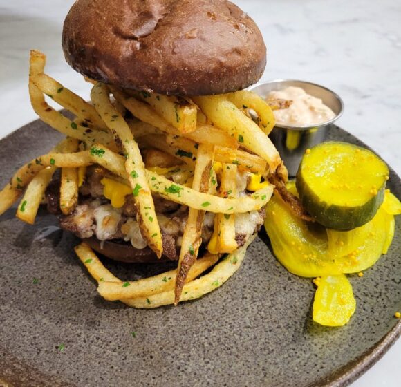 A hearty burger is topped with a generous portion of French fries and melted cheese, served on a round, speckled plate. On the side, there are pickle slices and a small metal cup filled with a creamy dipping sauce. The dish is arranged on a white marble surface. - Hausion