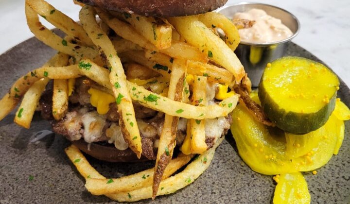 A hearty burger is topped with a generous portion of French fries and melted cheese, served on a round, speckled plate. On the side, there are pickle slices and a small metal cup filled with a creamy dipping sauce. The dish is arranged on a white marble surface. - Hausion