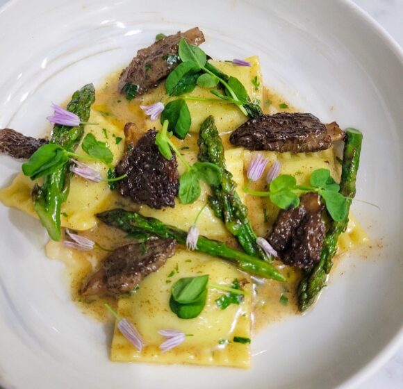 A white plate features a gourmet dish of ravioli garnished with morel mushrooms, asparagus spears, fresh herbs, and edible flowers in a light sauce. The presentation is artistic, showcasing a mix of textures and vibrant green and brown colors against the pale pasta. - Hausion