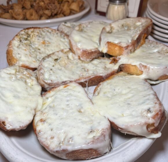 A plate of toasted bread slices generously topped with melted cheese. The bread appears crispy around the edges, and the cheese is golden and slightly bubbly. In the background, there is a glimpse of another dish and a stack of small white plates. - Hausion