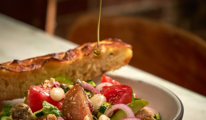 A close-up of a Mediterranean salad with tomatoes, tuna, onions, capers, and olives in a bowl on a table. A hand is drizzling olive oil over the salad, with a toasted slice of bread resting on the rim of the bowl, and a blurred background adding depth to the scene. - Hausion