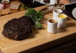 A charred steak rests on a wooden cutting board garnished with herbs and accompanied by three small white cups containing various sauces. A grazing board with assorted meats and cheeses, a glass of red wine, and a plate are visible in the background. - Hausion