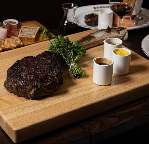 A charred steak rests on a wooden cutting board garnished with herbs and accompanied by three small white cups containing various sauces. A grazing board with assorted meats and cheeses, a glass of red wine, and a plate are visible in the background. - Hausion