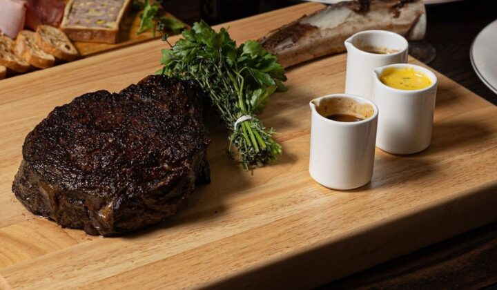 A charred steak rests on a wooden cutting board garnished with herbs and accompanied by three small white cups containing various sauces. A grazing board with assorted meats and cheeses, a glass of red wine, and a plate are visible in the background. - Hausion