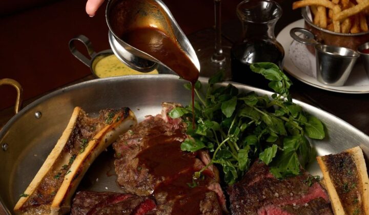 A hand pouring brown sauce over an assortment of grilled meats and bone marrow in a large pan, garnished with fresh greens. Surrounding the pan are a glass of red wine, a small jug of oil, a bowl of fries, and a silver gravy boat. - Hausion