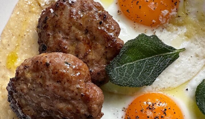 A close-up of a plate featuring two fried eggs with vibrant orange yolks, two seasoned sausage patties, fresh sage leaves, and a sprinkle of grated cheese on a creamy base. The dish is garnished with black pepper and a drizzle of olive oil. - Hausion